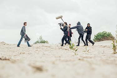 Man walking across sand towards film crew with cameras and microphones working on video production