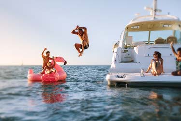 Young group hanging out on luxury yacht and inflatable flamingo raft at sunset, one man jumping off yacht into water